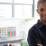 A woman smiling into the camera with her arms crossed in front of a desk with books on it.