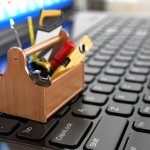 Close up of a very small toolbox on the the keyboard of a laptop filled with hammers, saws, levels, and other small tools.