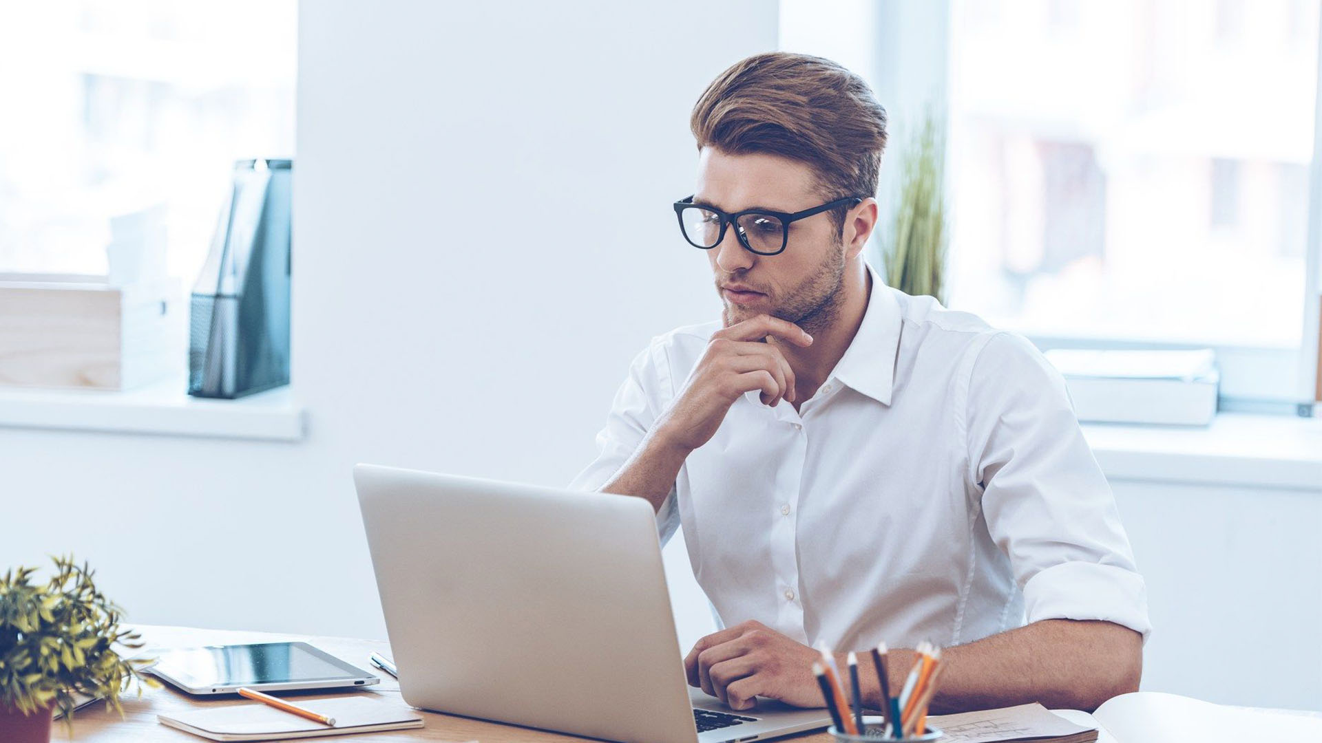 A man wearing glasses and a white button up shirt while working on his laptop at his desk.