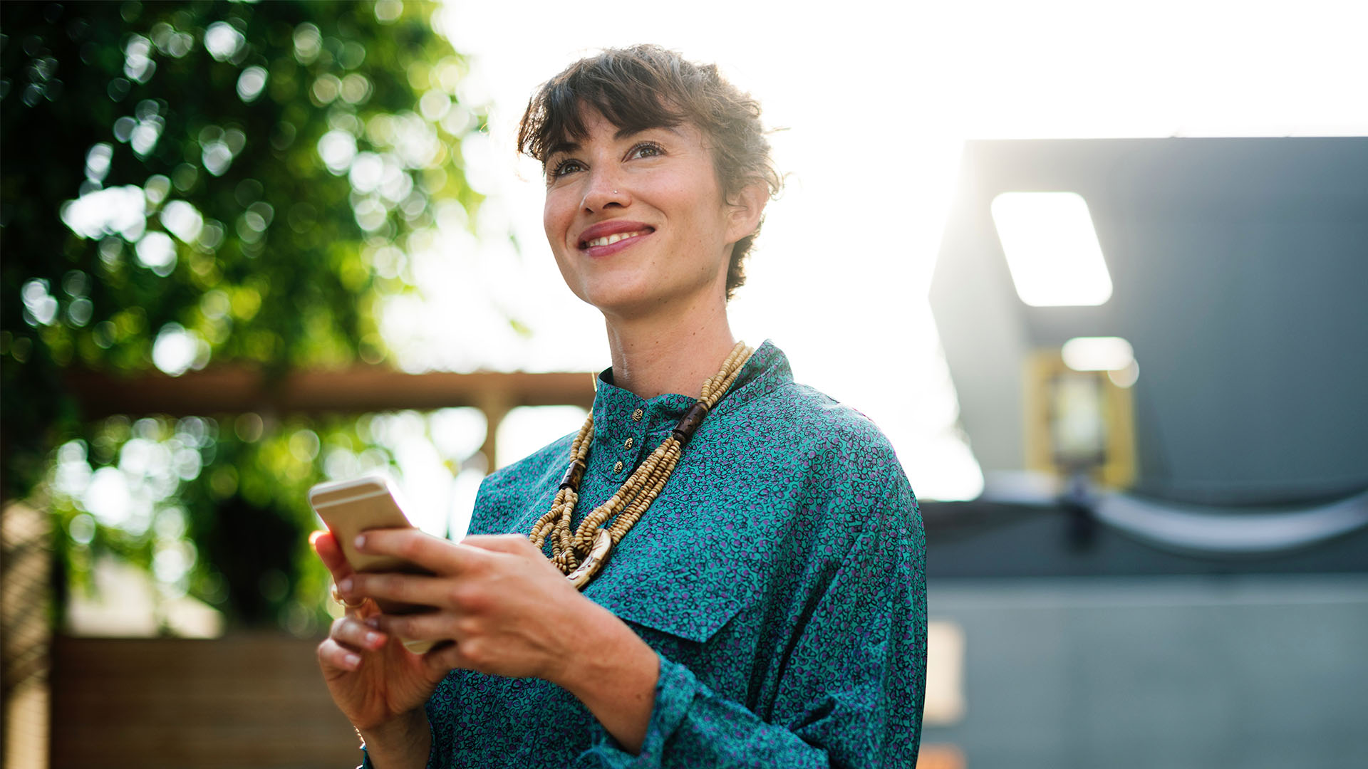 A woman outside on her smartphone looking up at the sky smiling on a sunny day with trees and buildings in the background.