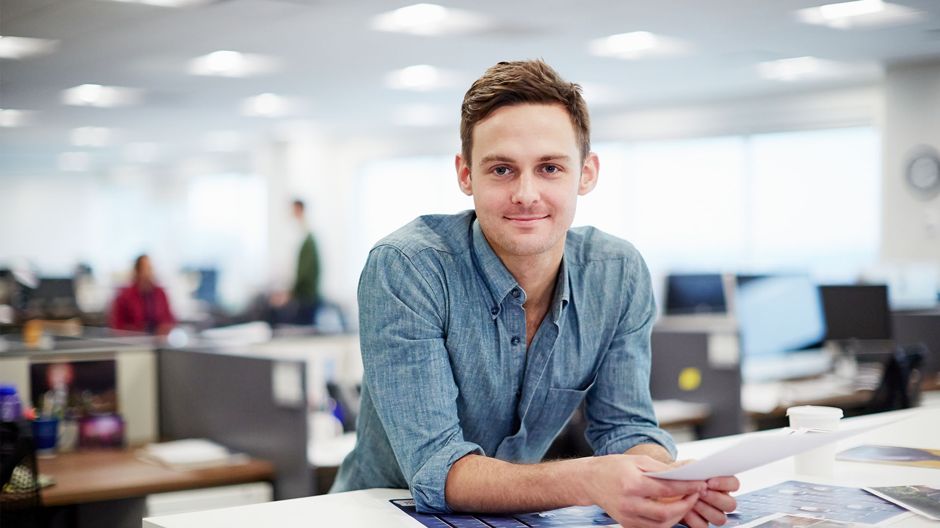 A man wearing a blue button up shirt smiling into the camera standing behind a desk in an open office space.