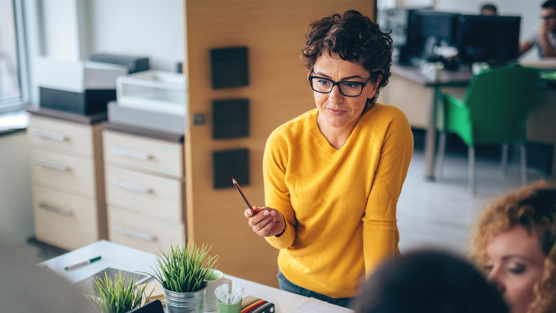 A woman standing up at a desk talking to other employees about project management while holding a pen.