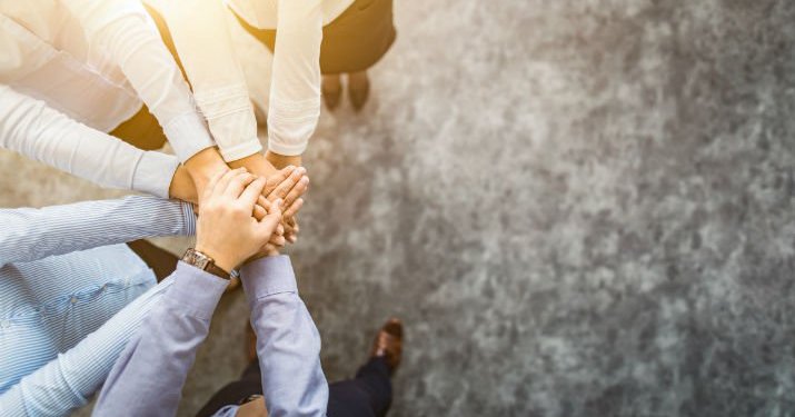 Overhead view of a group of people with their hands in the circle on top of each other as a representation of teamwork and globalization of HR leadership.