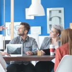 A modern workspace with vibrant blue walls and a group of co-workers sitting at a table looking up at a man standing up giving them instructions.