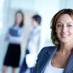 A business woman smiling into the camera with three other professionals out of focus in the background..