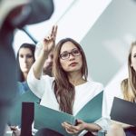 A small group of female HR students holding notebooks and listening to a lesson while one student is raising her hand.